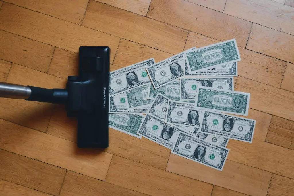 Vacuum cleaner head on a wooden floor appearing to suck up scattered one-dollar bills, symbolizing cash drain, poor expense control, or inefficient financial management.