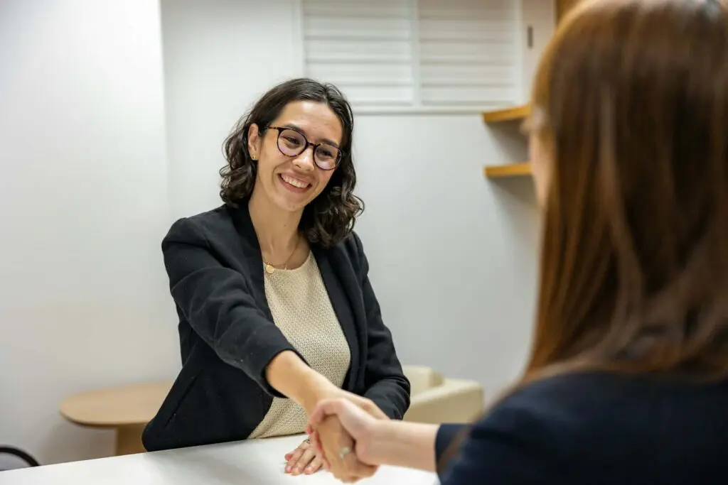 Smiling woman in business attire shakes hands with another person across a desk, likely during a job interview or meeting.
