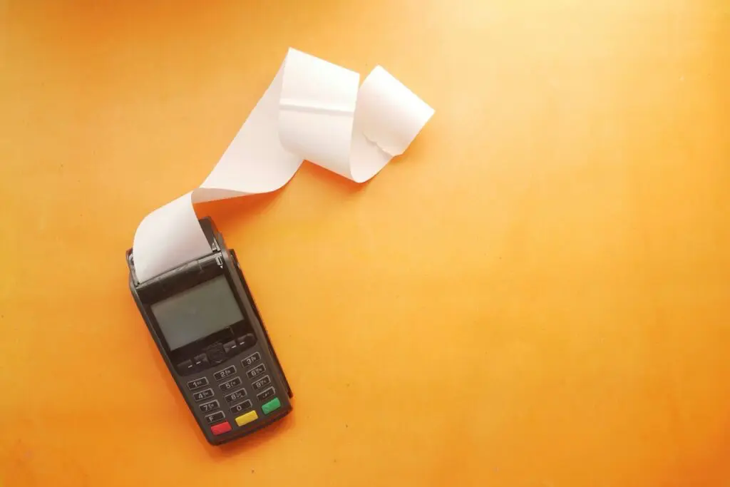 The image shows a payment terminal (POS machine) with a long paper receipt curling out of it, placed on an orange surface.