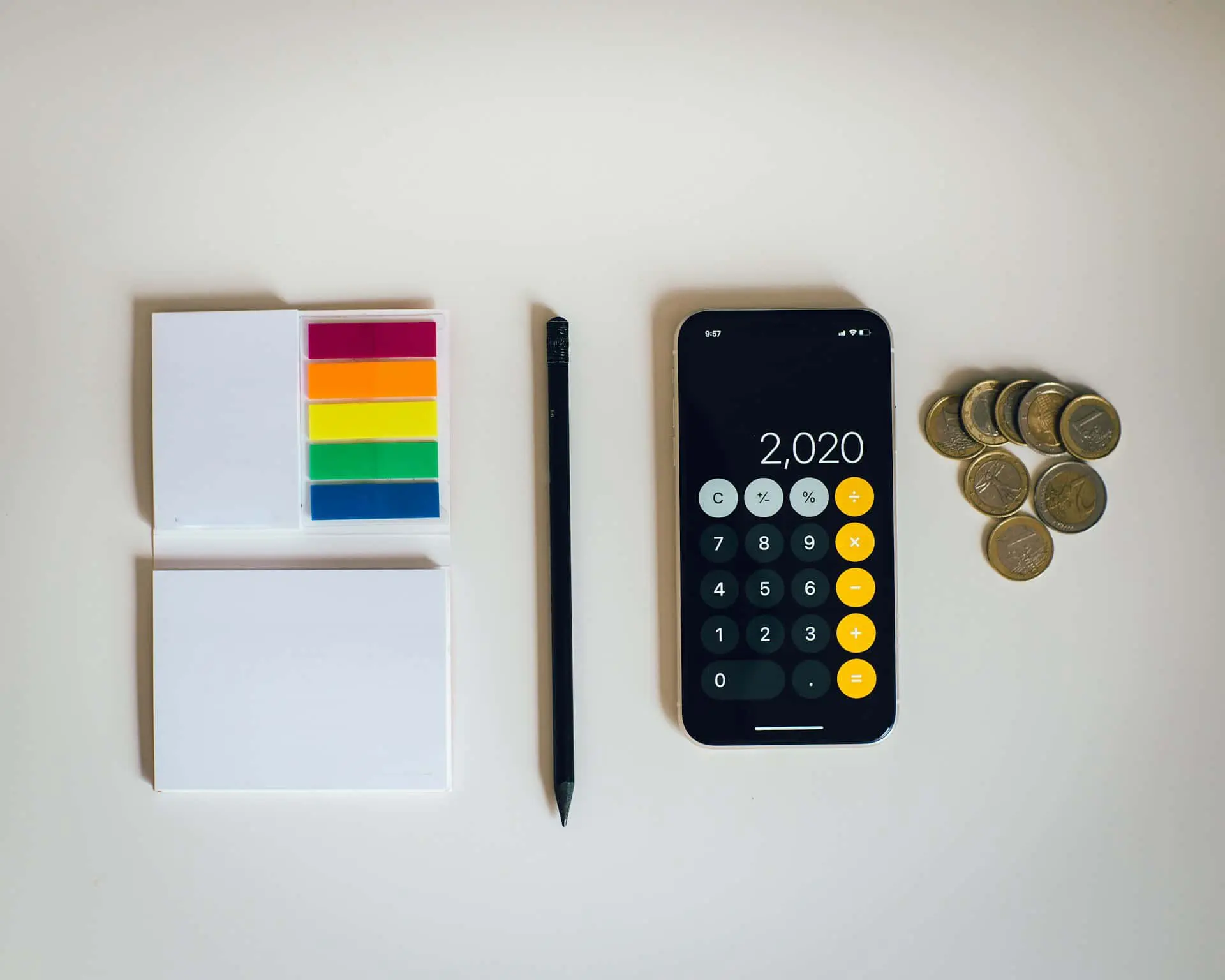 Flat lay of financial planning tools including sticky notes with colorful tabs, a pencil, a smartphone calculator displaying “2,020,” and euro coins, all arranged on a clean white surface.