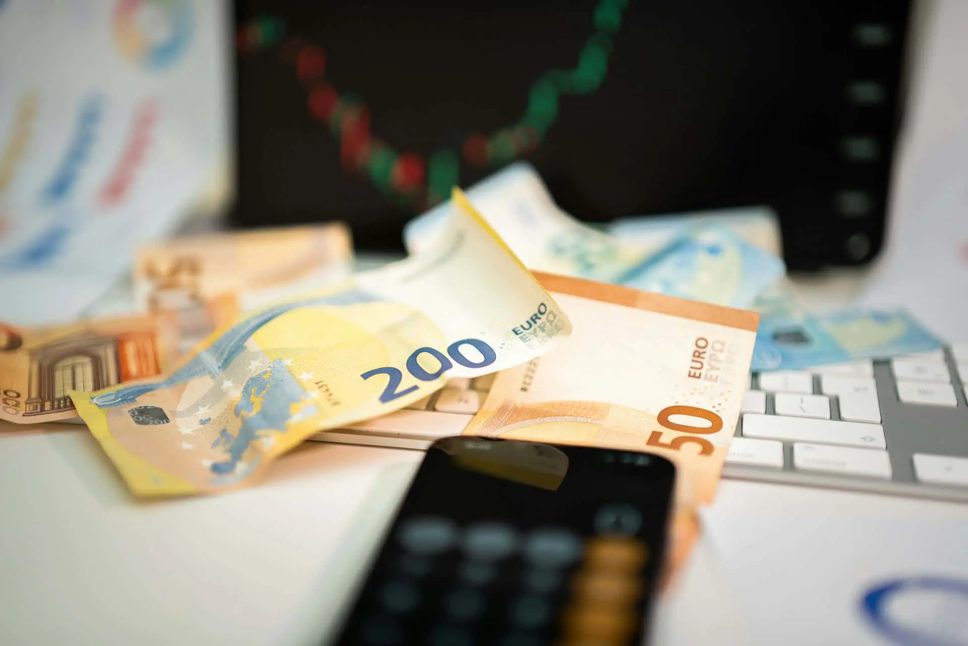 Close-up image of various euro banknotes (including €200, €50, and €20) scattered on a desk. The background includes part of a keyboard, a blurred smartphone screen with a calculator app open, and a laptop screen showing a green and red candlestick chart, indicating financial data or stock market activity. The image conveys a theme of financial planning or investment.