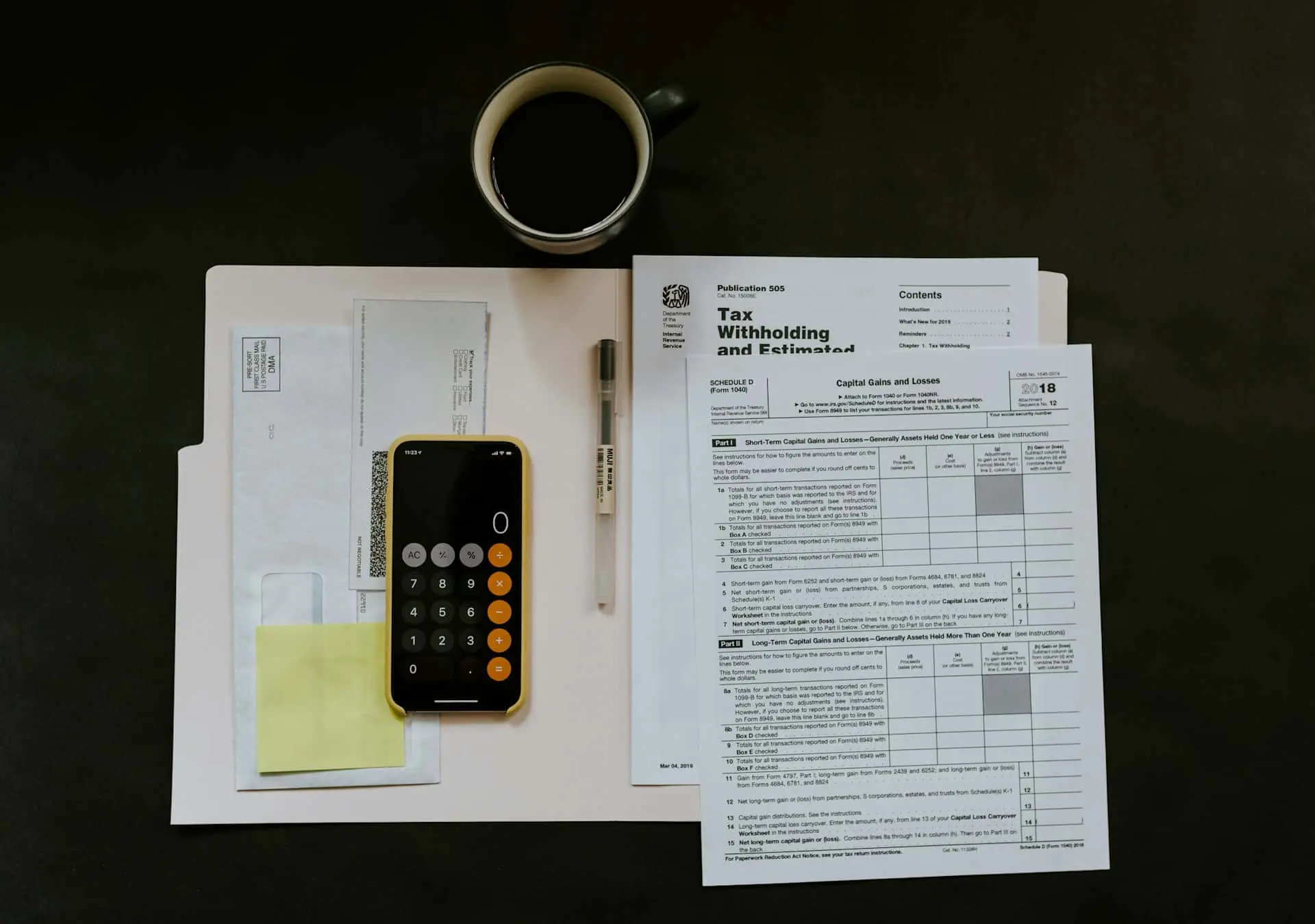 A flat lay of tax preparation materials on a dark surface. The image includes U.S. tax documents (such as IRS Form 1040 Schedule D for capital gains and losses), a pink file folder, envelopes, a smartphone displaying a calculator app with a zero balance, a pen, a sticky note pad, and a black coffee mug. The setup conveys a tax filing or financial planning context.