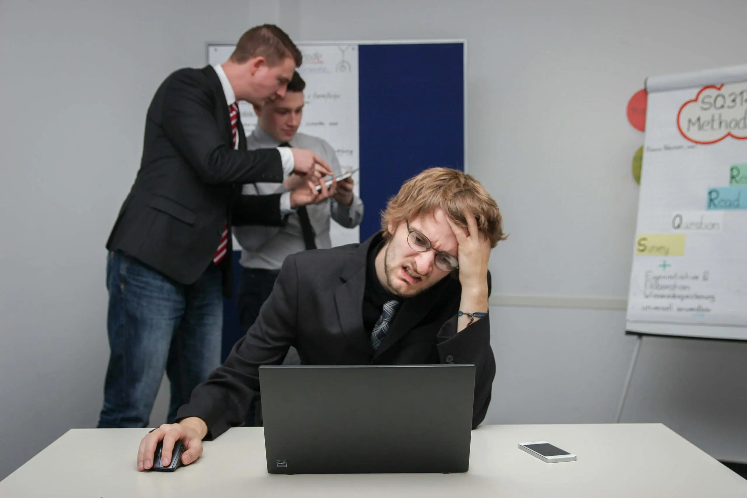 Frustrated office worker grimacing at a laptop while colleagues discuss in the background, symbolizing bloated enterprise software built for committees.