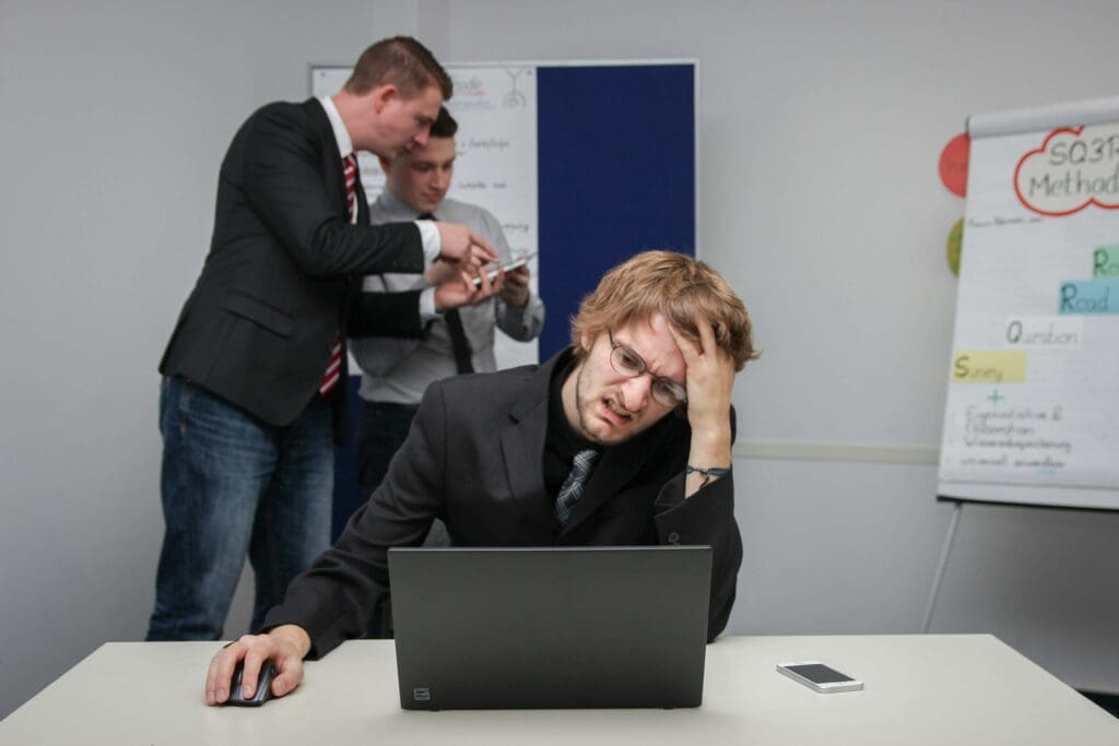 Frustrated office worker grimacing at a laptop while colleagues discuss in the background, symbolizing hard capex planning